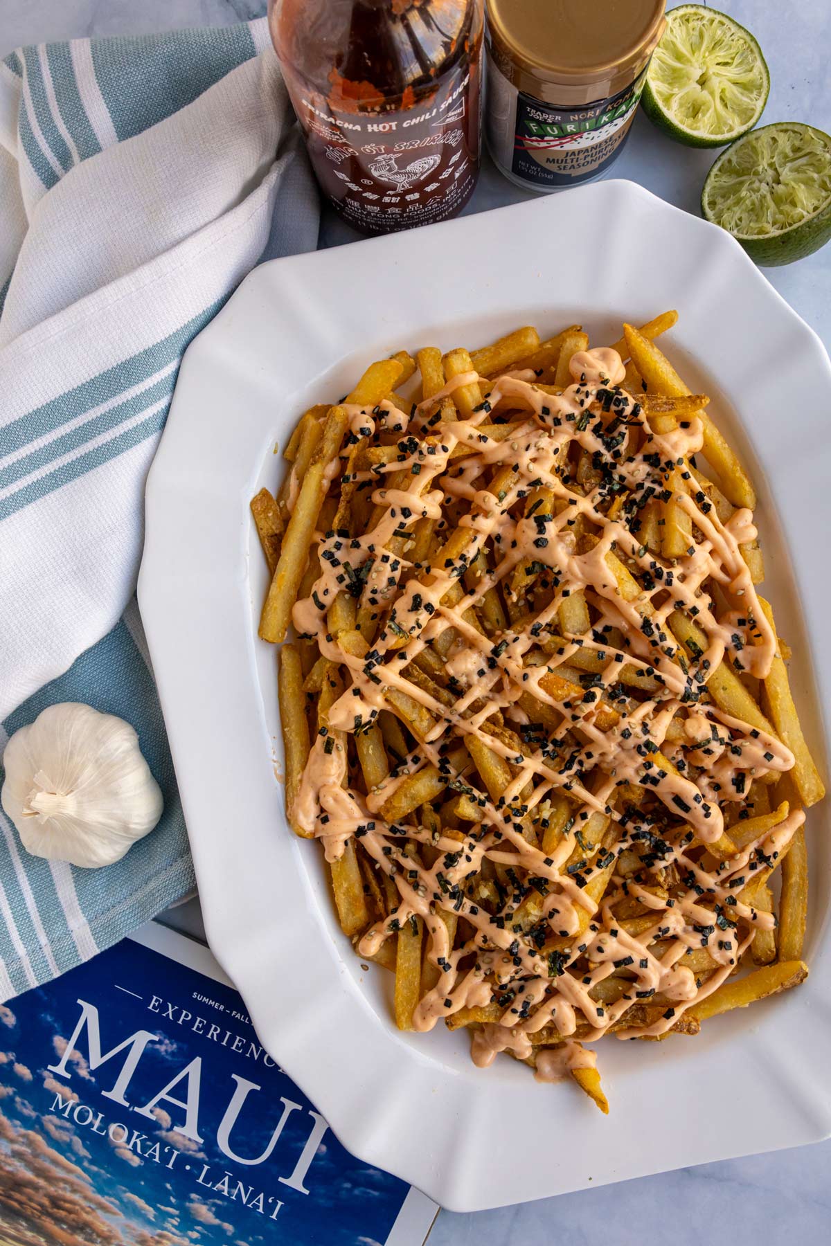 A platter of volcano fries with bottles of sriracha and furikake and a Maui magazine beside it.