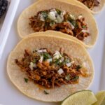 Braised and shredded chicken tacos on corn tortillas served on a white platter.
