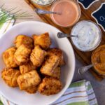 Kibbeling in a serving dish next to a platter of dipping sauces in small bowls.