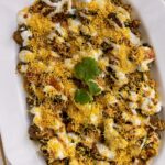 Aloo chana chaat in a white oval serving dish topped with yogurt, tamarind chutney, and sev.