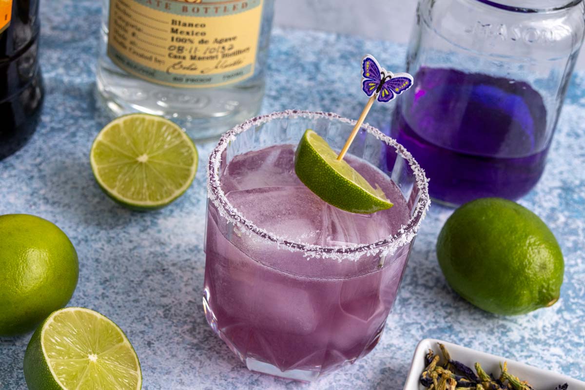 A butterfly pea flower margarita on a blueish surface with limes around it and bottles behind it.