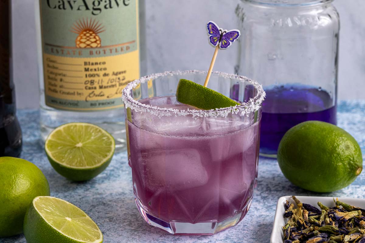 A butterfly pea flower margarita on a blueish surface with limes around it and bottles behind it.