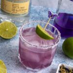 A butterfly pea flower margarita garnished with a purple butterfly skewer through a lime wedge.