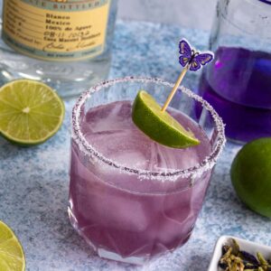 A butterfly pea flower margarita garnished with a purple butterfly skewer through a lime wedge.