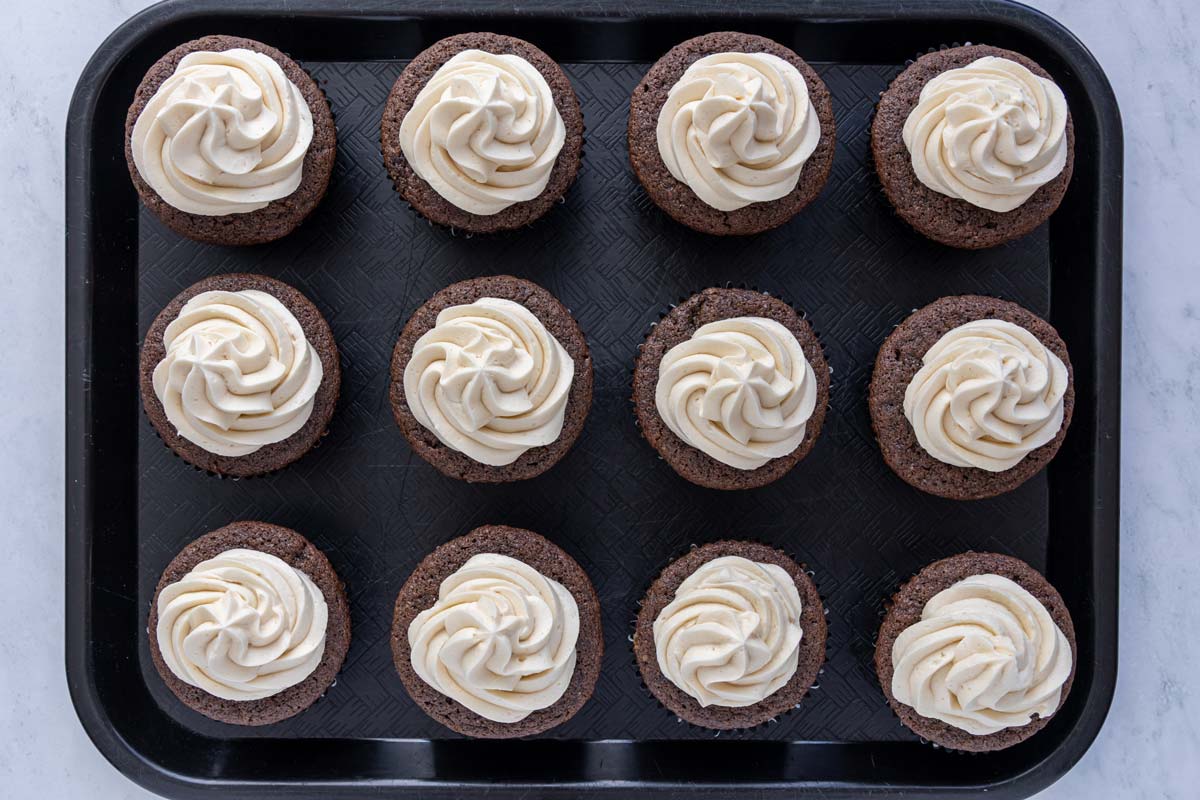 Bailey's frosting piped into swirls atop chocolate cupcakes on a black cafeteria tray.