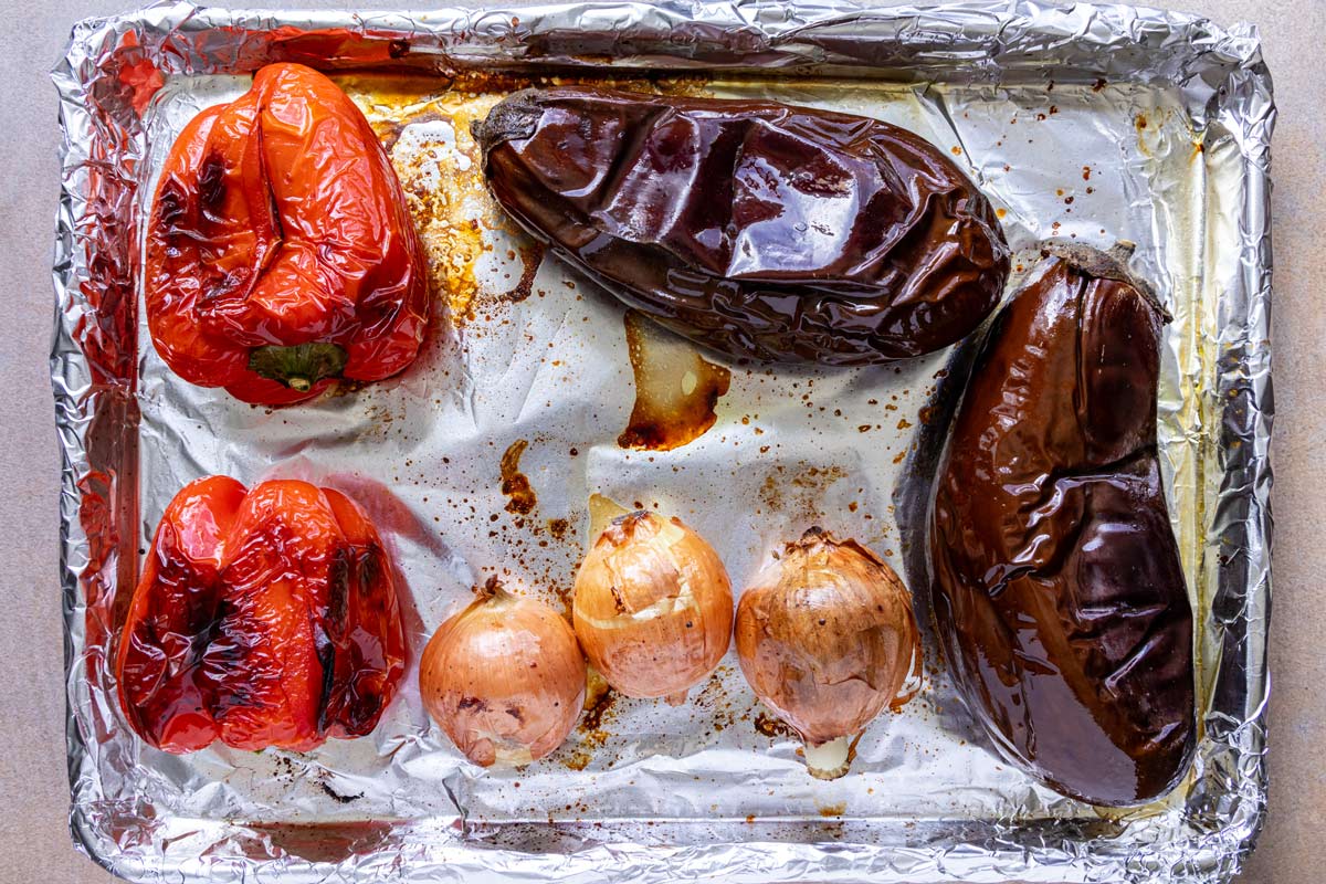 Eggplants, red peppers, and onions coated with olive oil on a foil lined sheet pan after roasting.