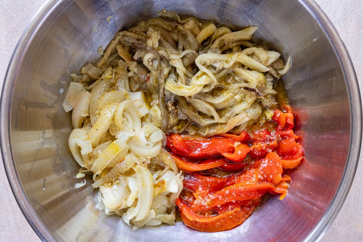 Strips of roasted eggplant, red peppers, and onion in a large metal mixing bowl topped with vinaigrette.
