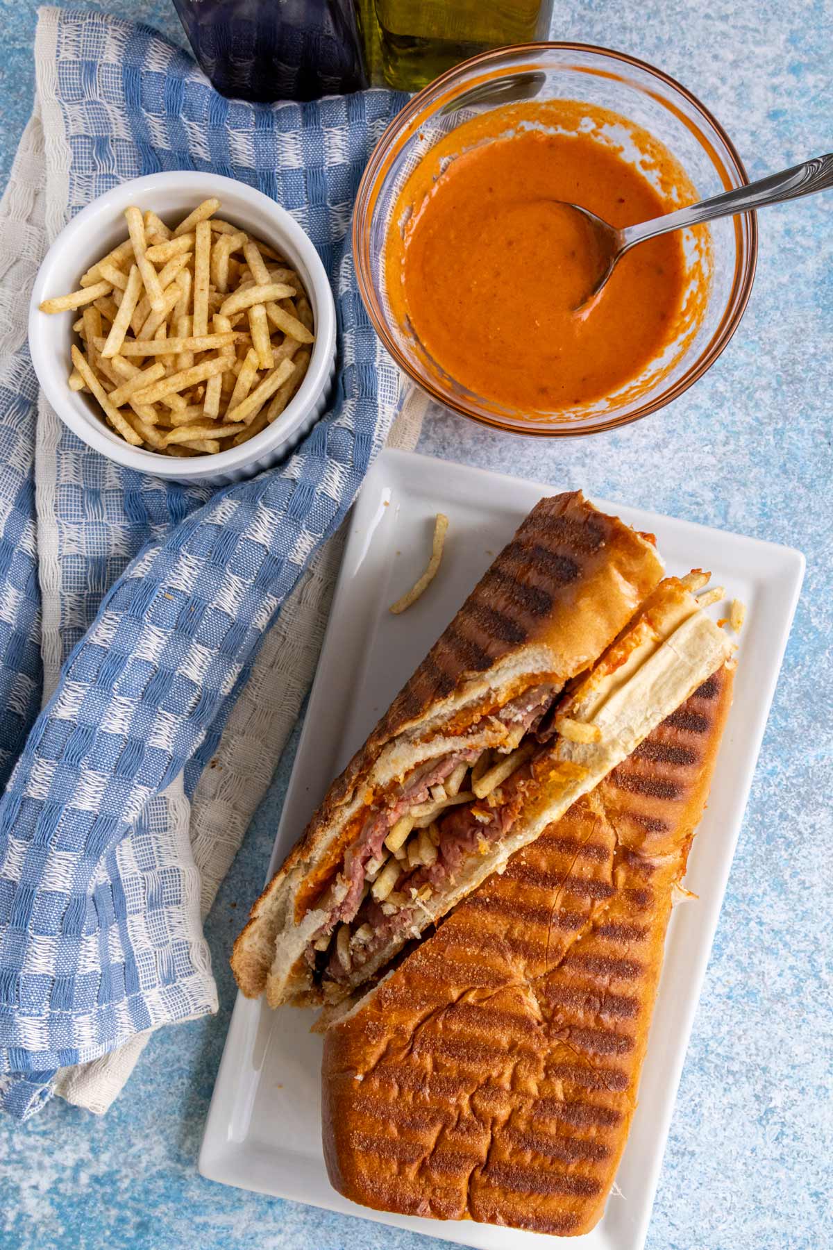 A pan con bistec Cuban steak sandwich with bowls of mojo rojo and potato sticks next to it.