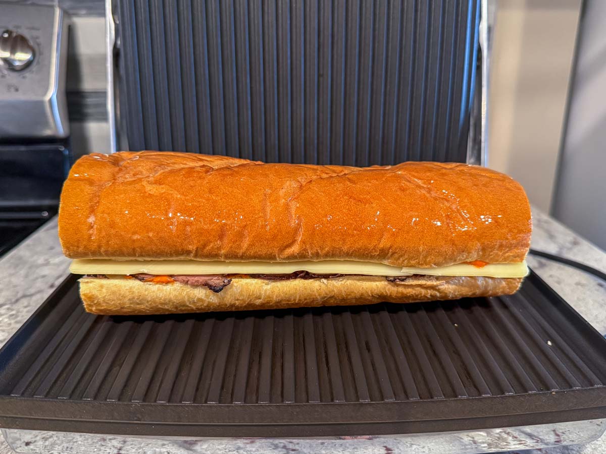 An assembled pan con bistec sandwich placed in a panini press before cooking.