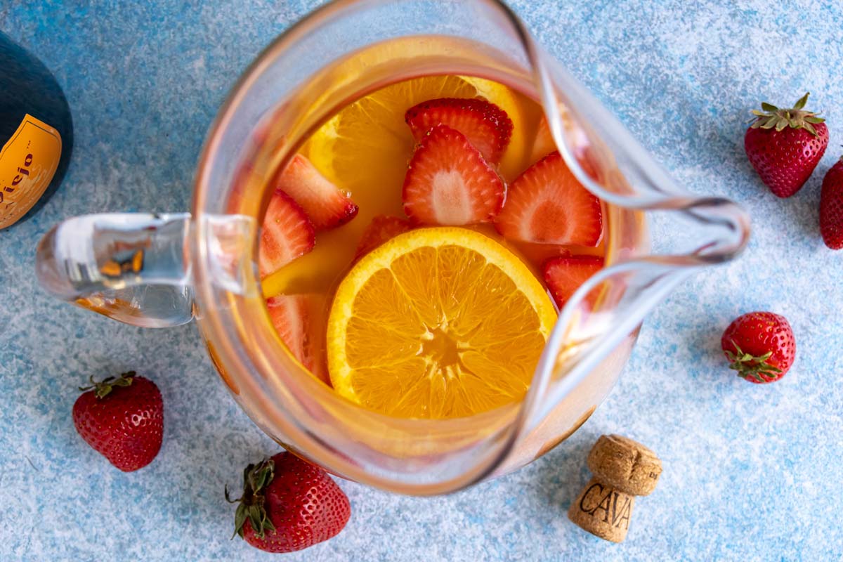 A glass pitcher of cava sangria on a bluish surface surrounded by strawberries.