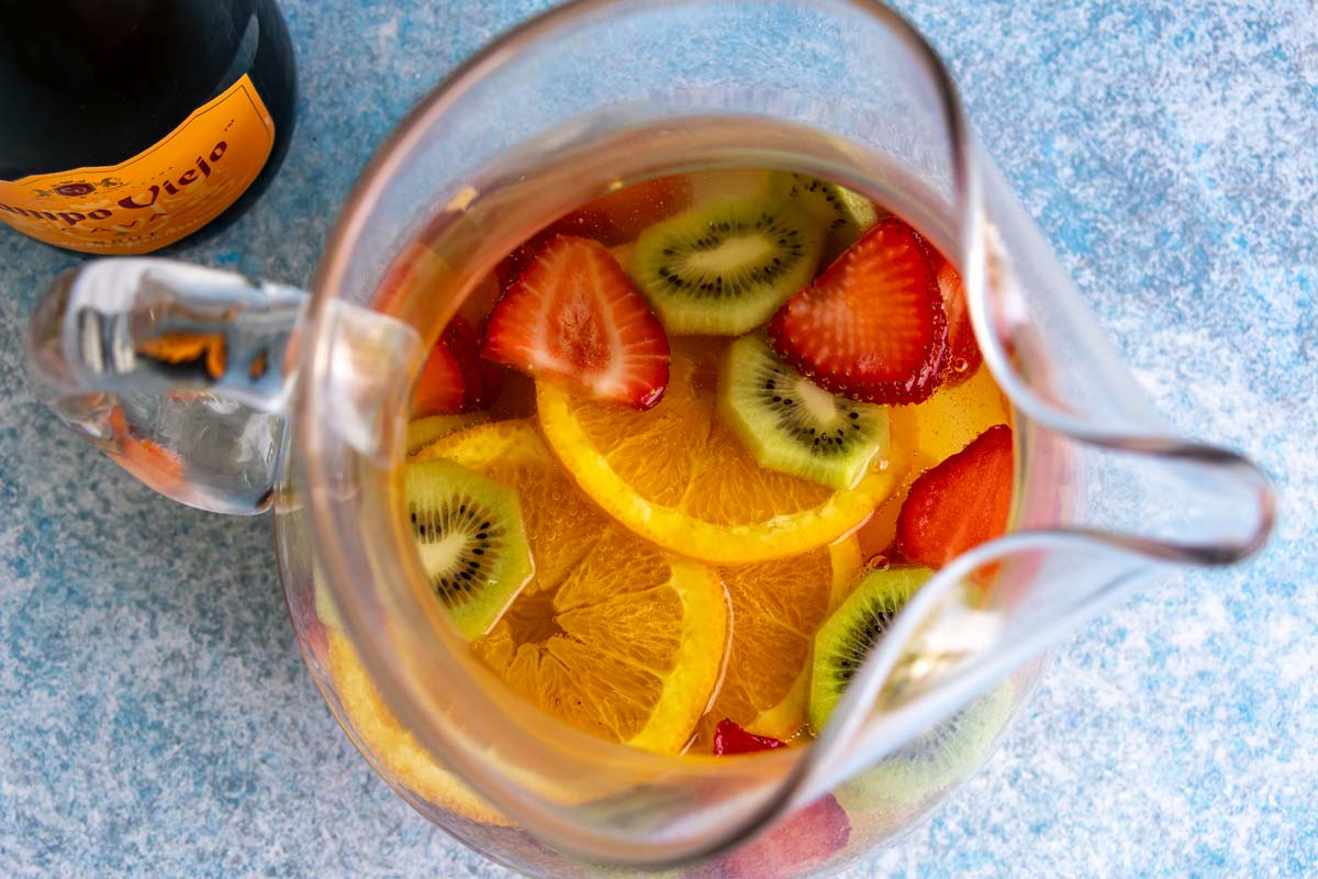 Cava sangria with sliced oranges, kiwi, and strawberries in a glass pitcher.