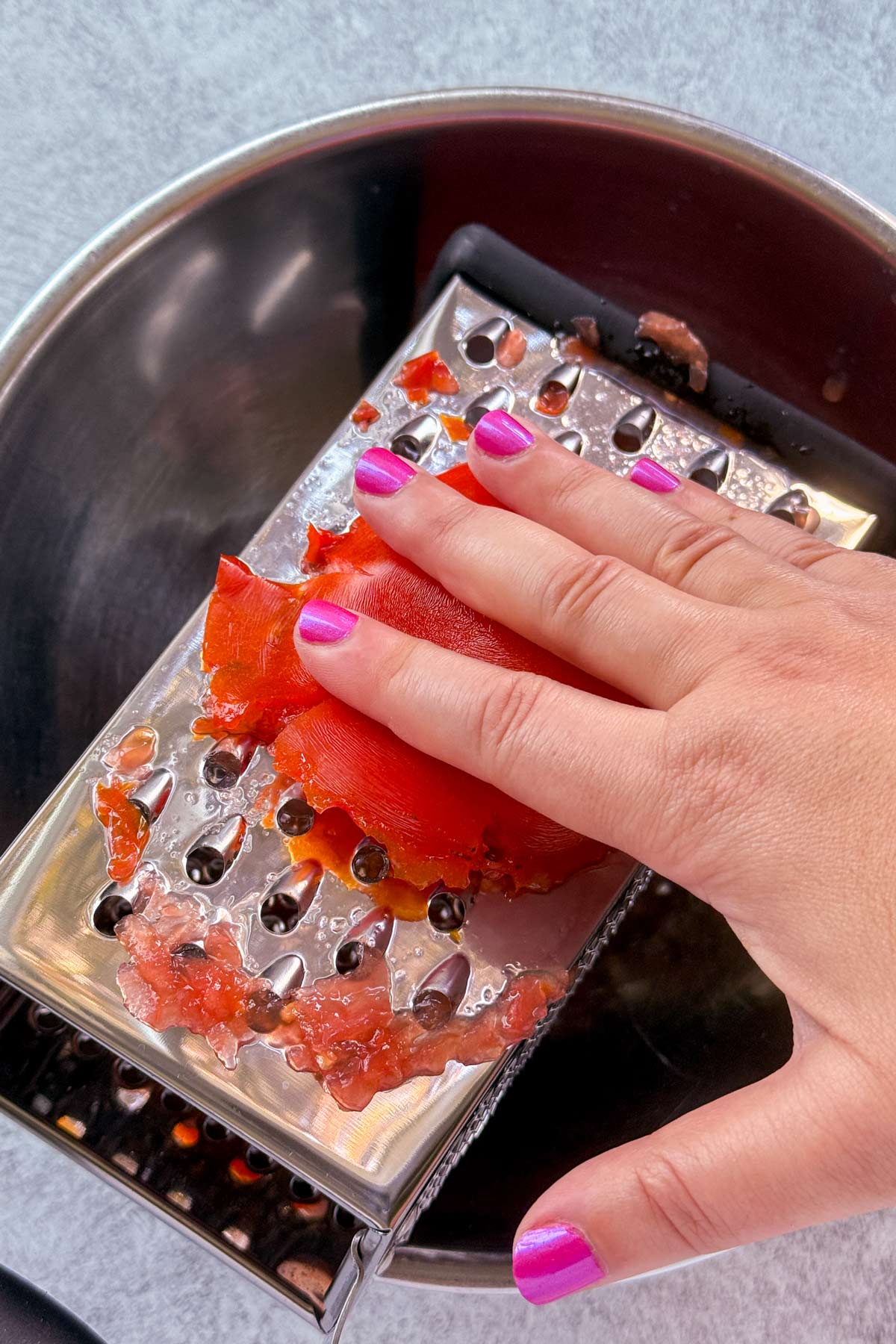 Grating half a ripe tomato on the side of a box grater set over a bowl.
