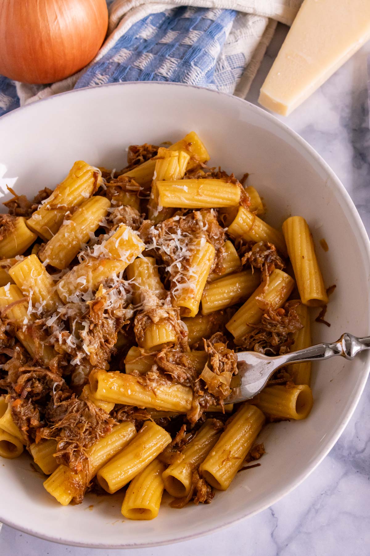A fork scooping up some rigatoni with Genovese sauce out of a shallow pasta bowl.