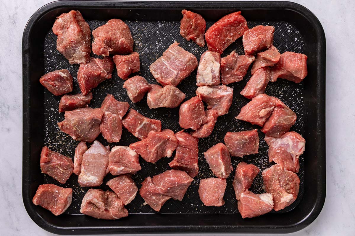 Cubes of chuck roast seasoned with salt and pepper on a black plastic cafeteria tray.