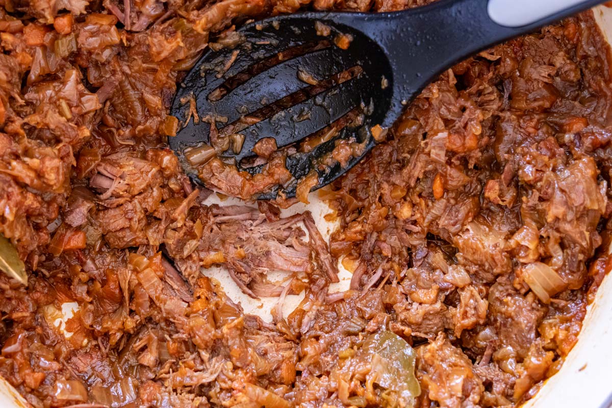 Using the back of a slotted spoon to break apart pieces of stewed beef into small shreds.