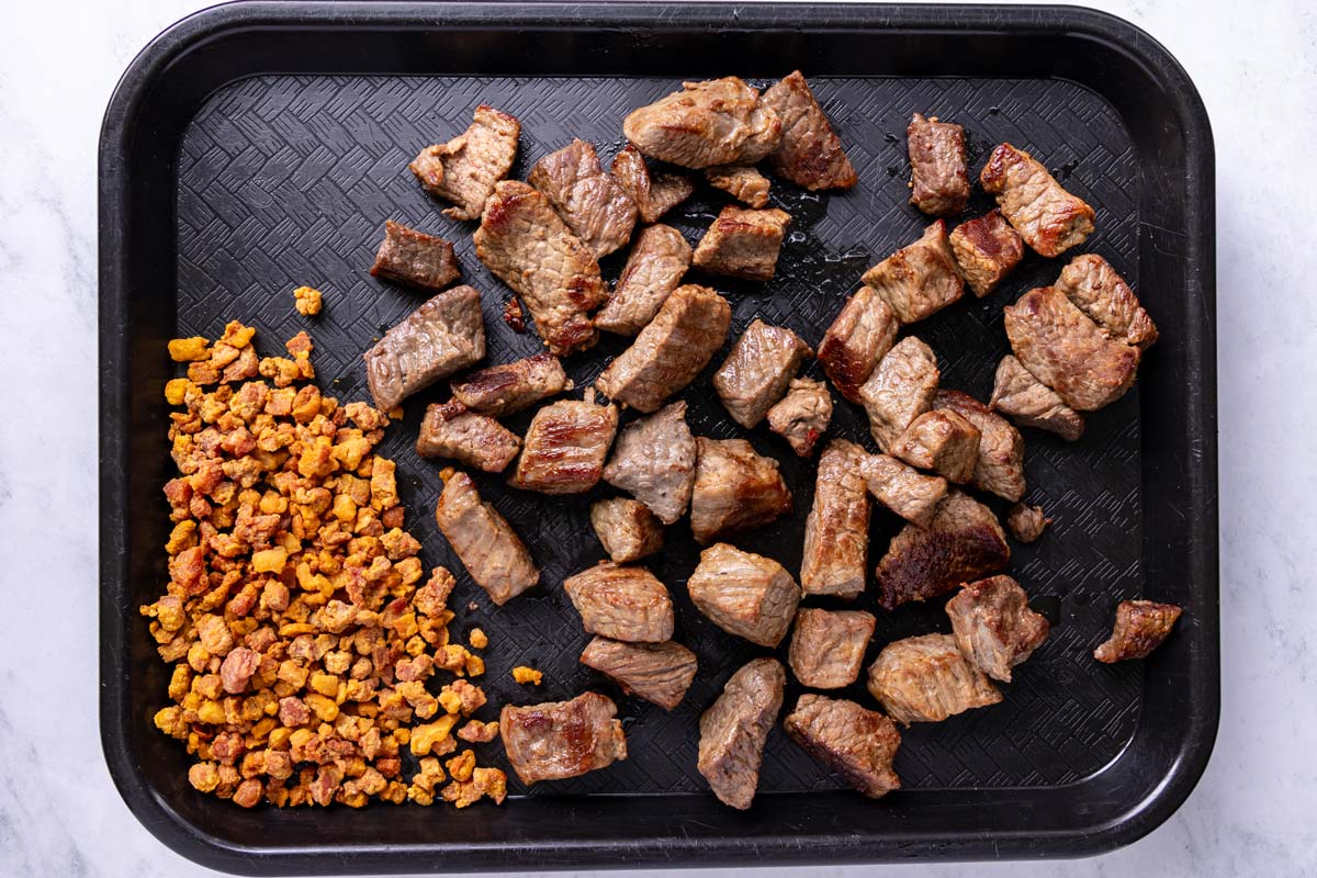 Seared beef cubes and diced pancetta on a black plastic cafeteria tray.