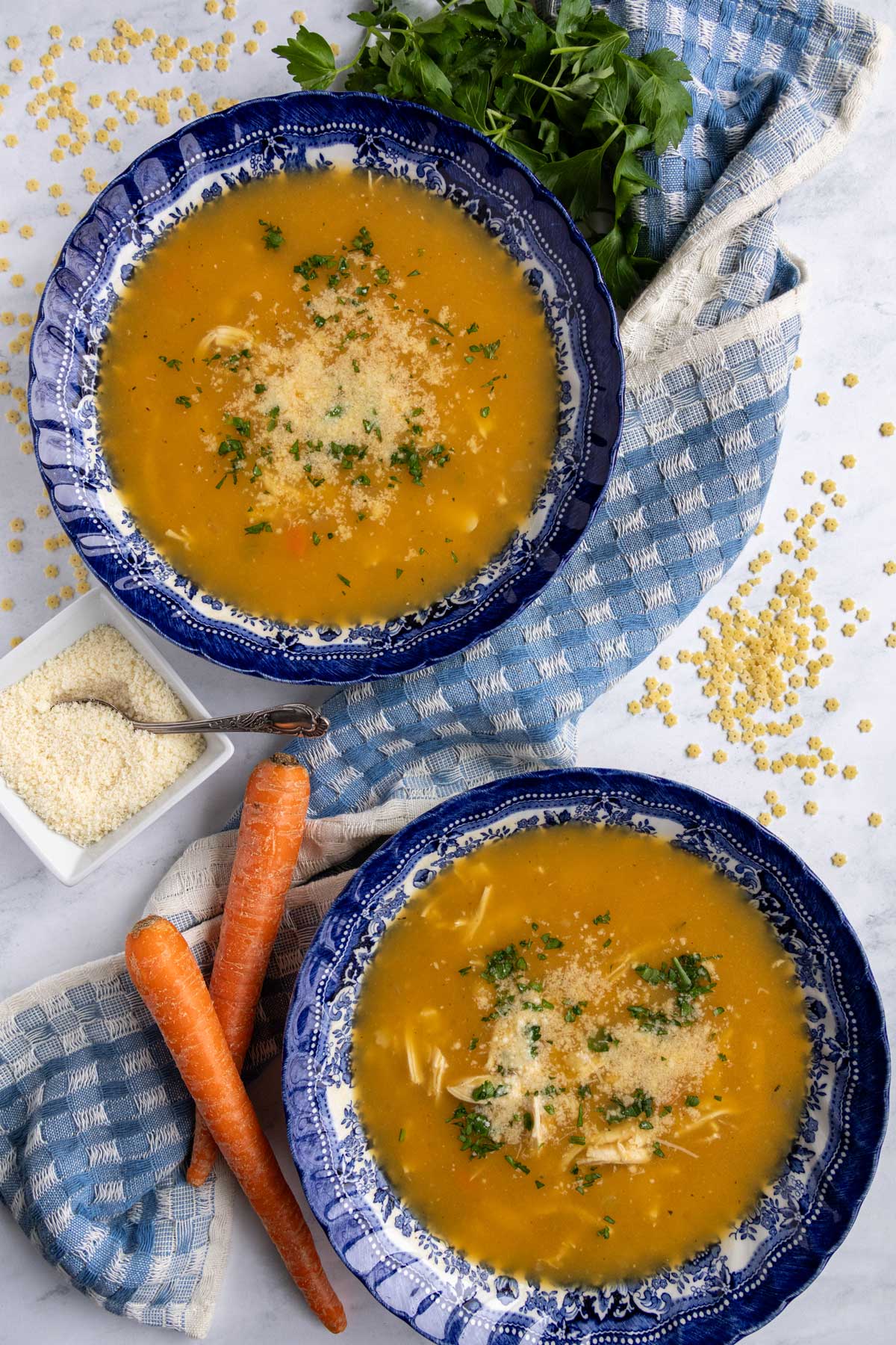 Two bowls of Italian chicken pastina soup with a blue gingham towel and a bowl of parmesan.