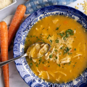 A spoon scooping some chicken pastina soup out of a blue and white shallow bowl.