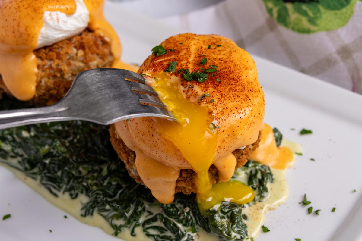 A fork piercing into the poached egg in a serving of Eggs Sardou, with runny yolk oozing out.