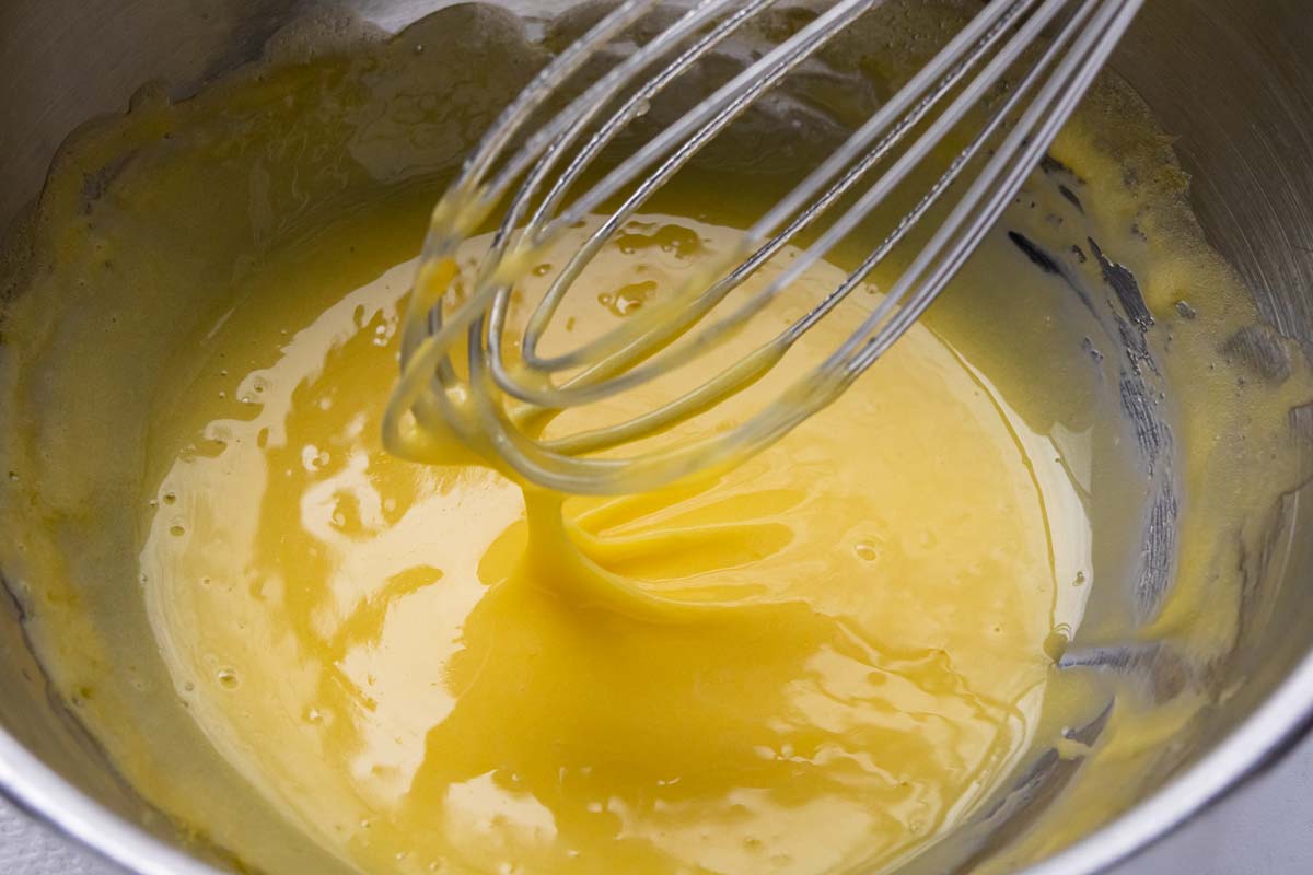 Hollandaise sauce dripping off a whisk lifting out of a metal mixing bowl.