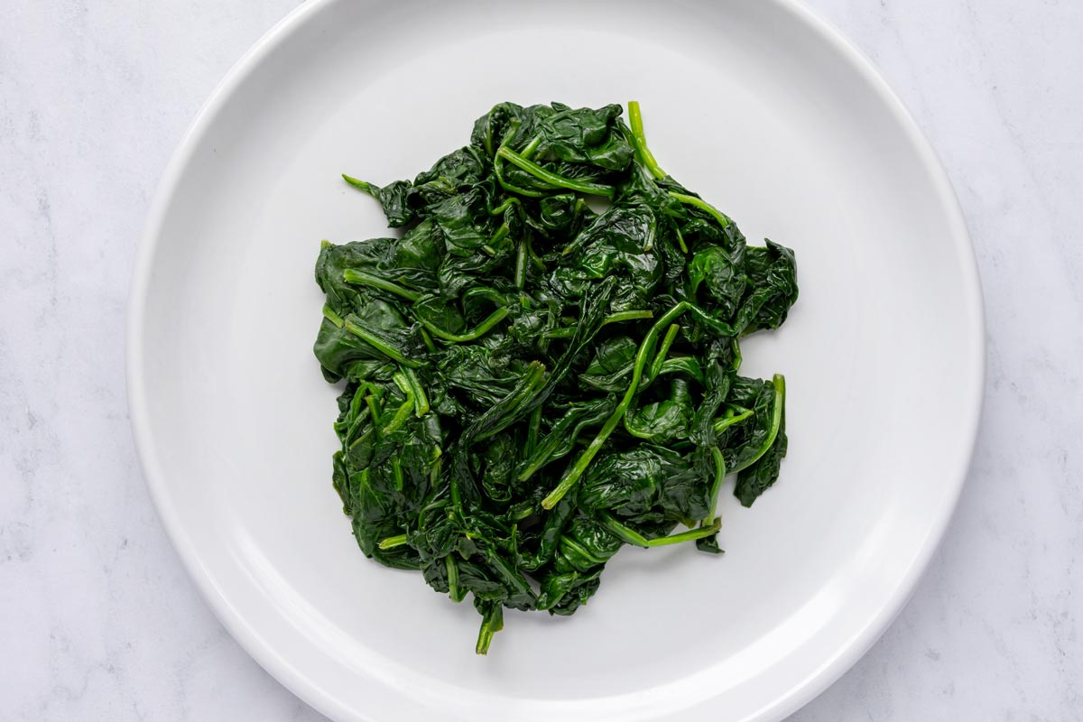 Wilted baby spinach leaves on a white plate after being squeezed of extra liquid.