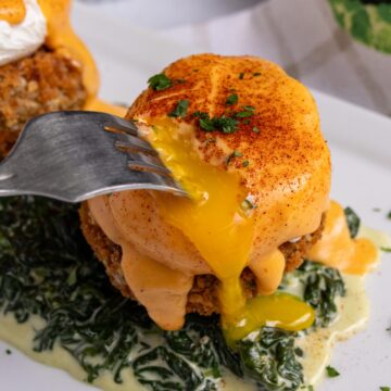 A fork piercing into the poached egg in a serving of Eggs Sardou, with runny yolk oozing out.
