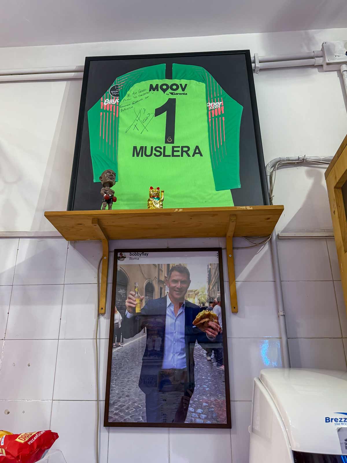 A Muslera jersey hanging above a framed photo of Bobby Flay holding a sandwich and drink.