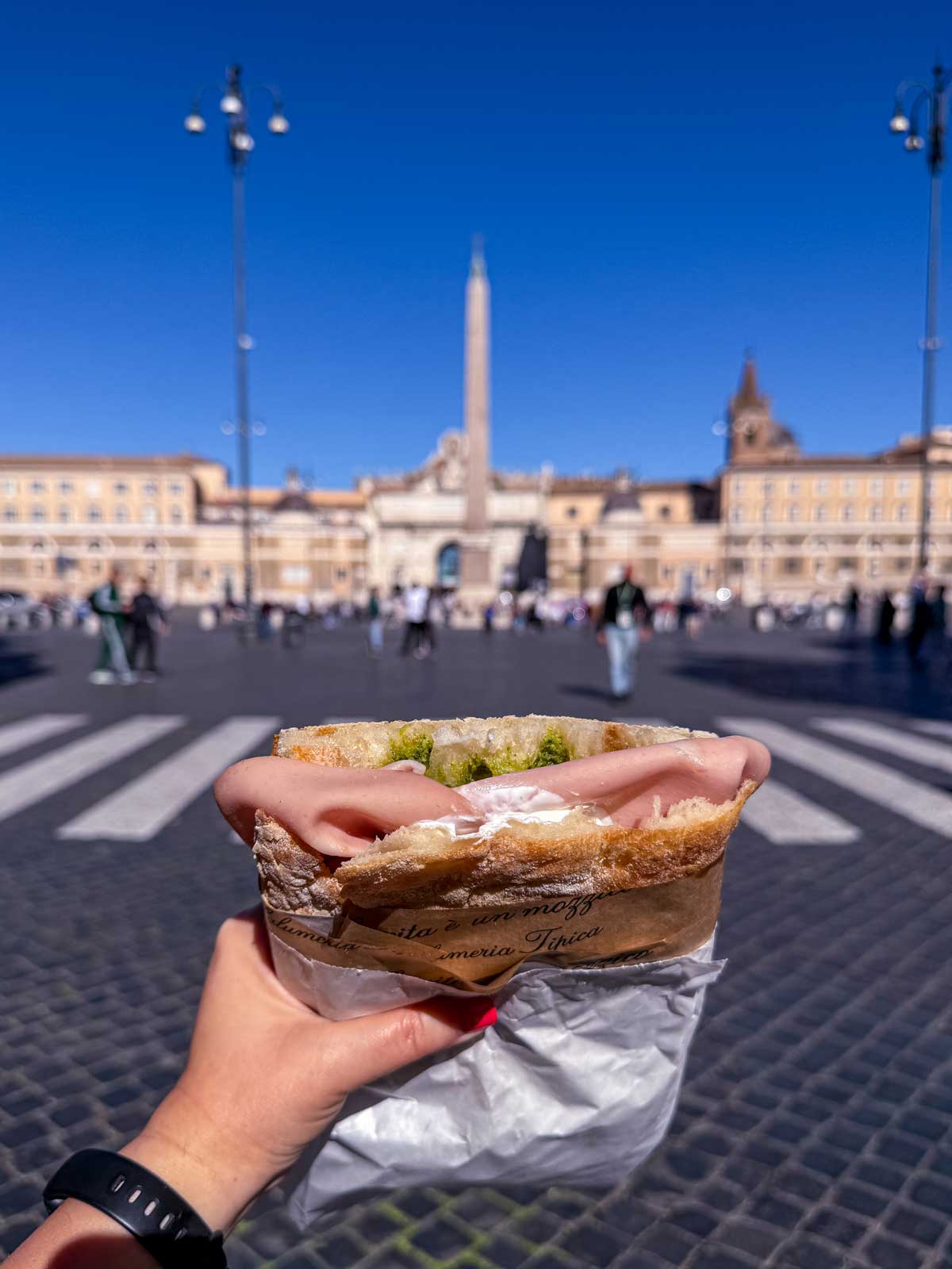 A hand holding a mortadella sandwich in Piazza del Popolo in Rome.