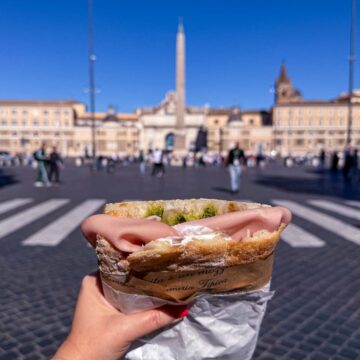 A hand holding a mortadella sandwich in Piazza del Popolo in Rome.