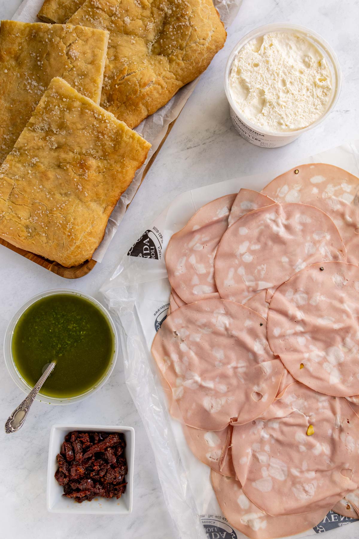 Ingredients for mortadella sandwiches on a white marble background.