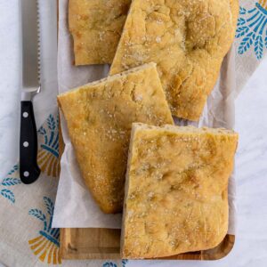 Four rectangular pieces of pizza bianca on a wooden platter with a serrated knife to the left.