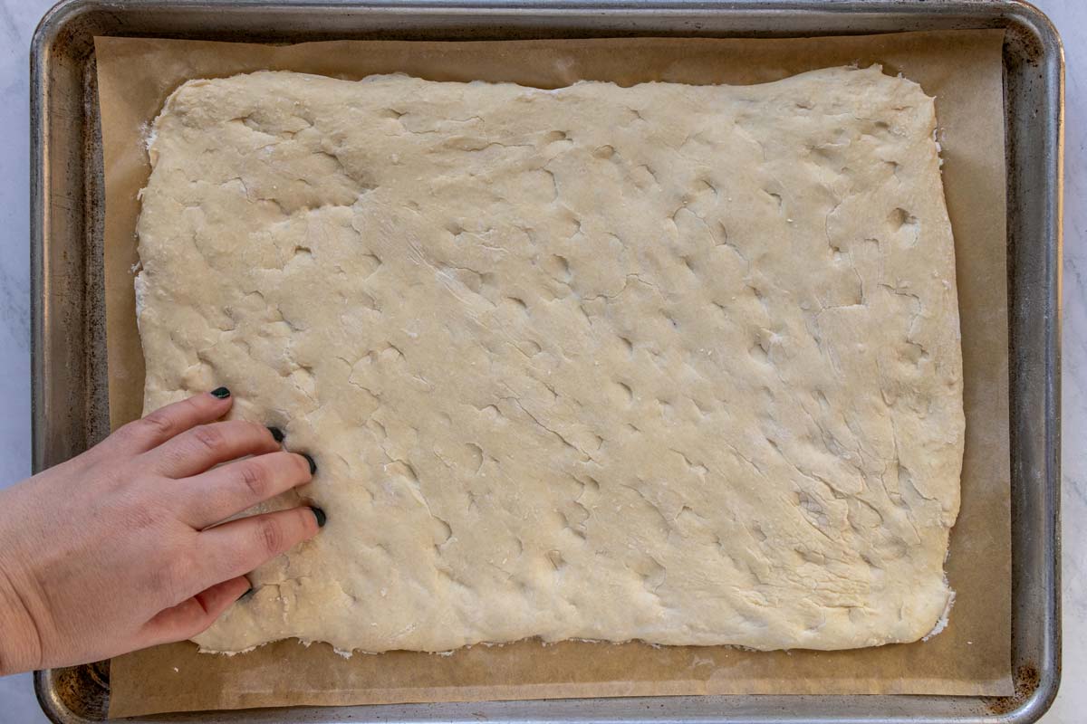 Fingers dimpling the top of a rectangular dough on a baking sheet.