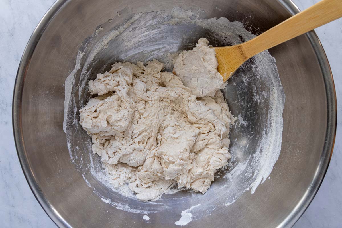 A partially mixed dough with a wooden spoon in a metal bowl.