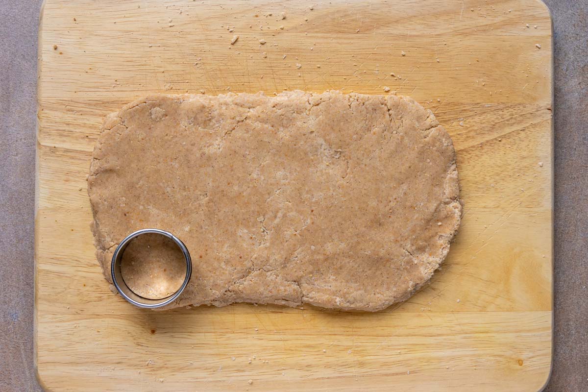 A flattened oval of dough on a wooden surface with a round cutter pressed into one corner.
