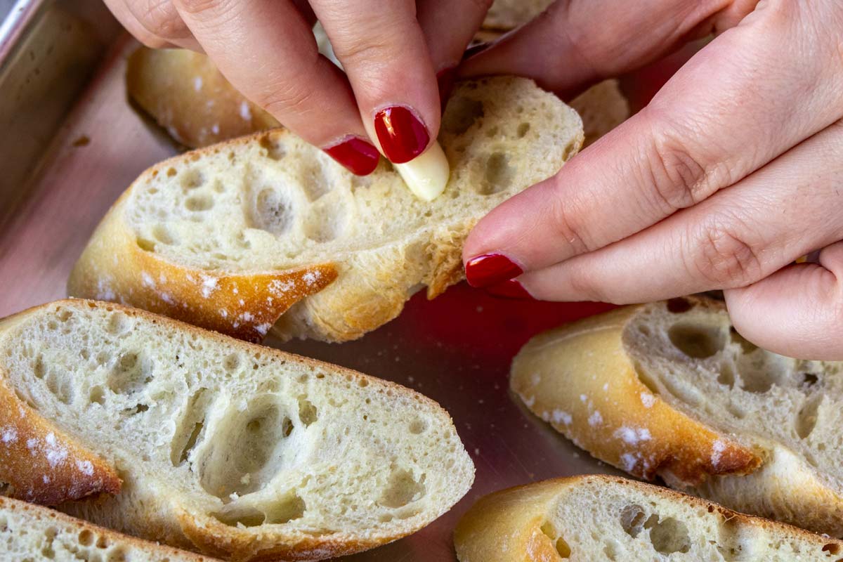 A hand rubbing a whole clove of garlic on lightly toasted slices of baguette.