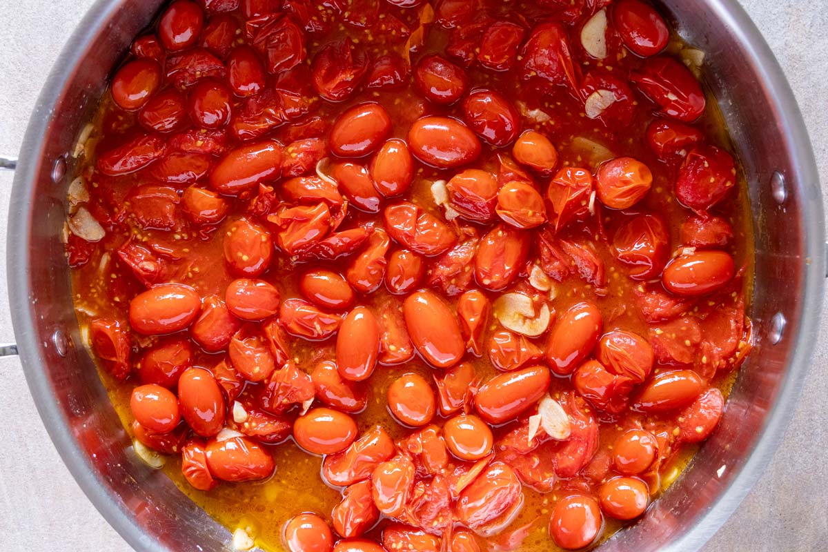 A mixture of whole and smashed cooked grape tomatoes in a wide skillet with garlic and olive oil.