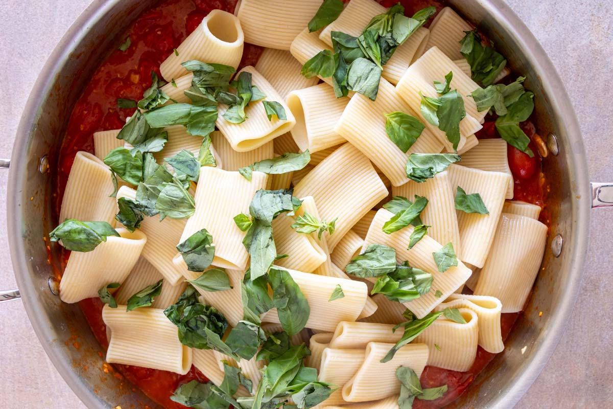 Boiled paccheri pasta and torn fresh basil added to a wide deep skillet of tomato sauce.