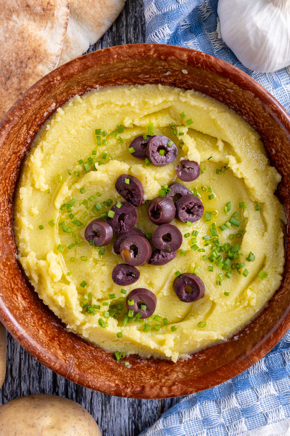 Greek garlic and potato spread (skordalia) topped with olives and chives in a brown bowl.