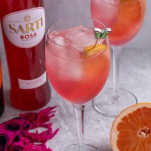 Two glasses of Sarti Spritz garnished with grapefruit slices, and a bottle of Sarti Rosa.