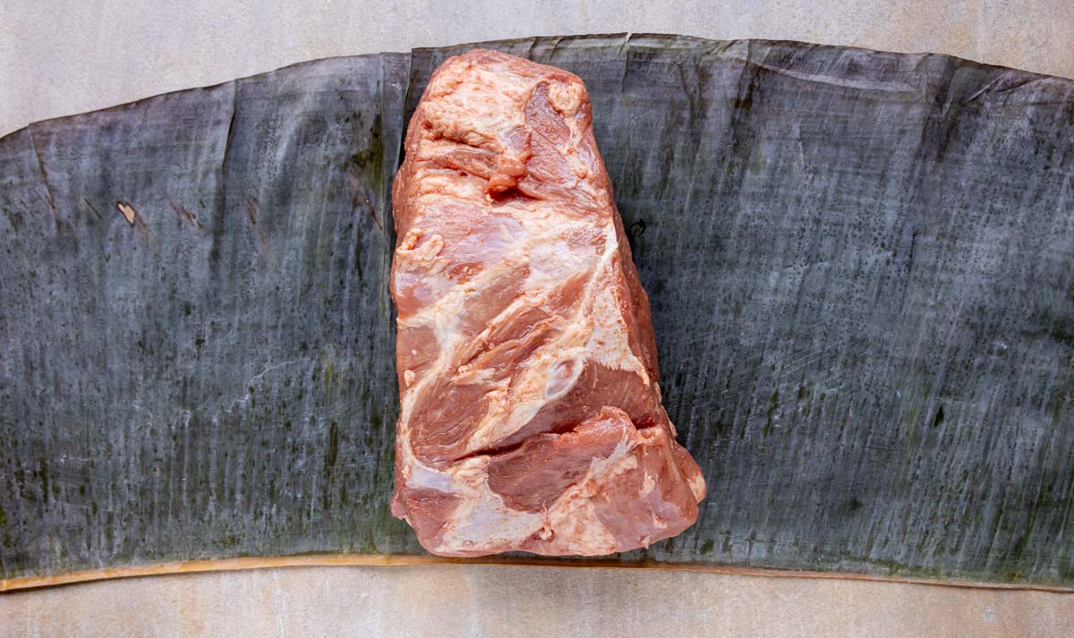 A trimmed bone-in pork butt seasoned and placed on a fresh banana leaf.
