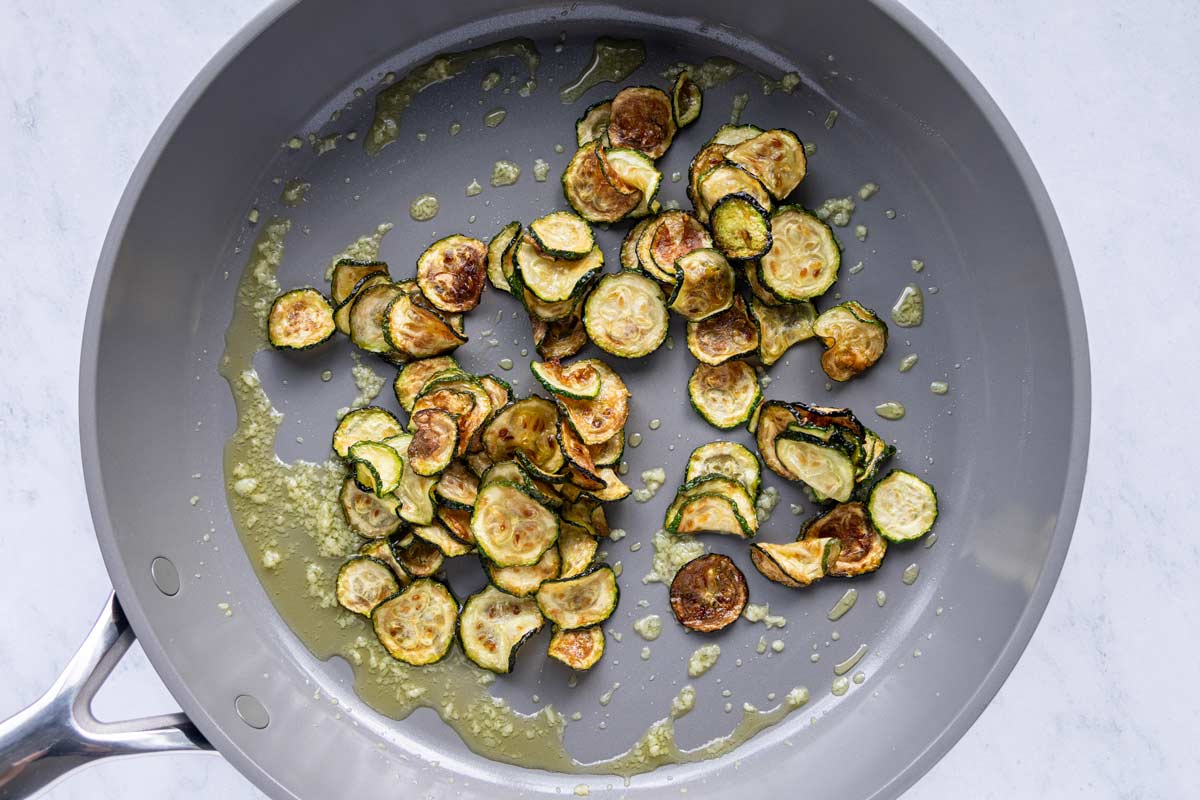 Fried zucchini coins added to a skillet with olive oil and garlic.